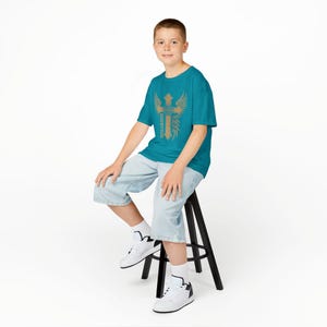 May include: Teal t-shirt featuring a gold cross design with wings and the word "BLUEPTION" vertically printed on the side. The person is wearing light blue denim shorts and white sneakers with black accents. The person is sitting on a black stool.