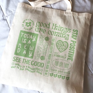 May include: A white canvas tote bag with green text and graphics. The text includes "good things are coming", "stay positive", "today is a new day", "see the good", "you got this", and "know your worth".