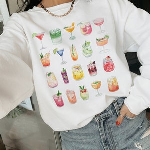 May include: White sweatshirt with a colorful watercolor design of various cocktails and drinks. The sweatshirt has a crew neck and long sleeves.