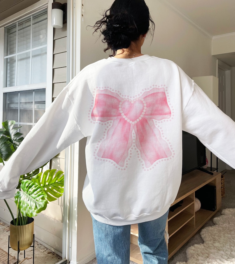 May include: White sweatshirt with a pink watercolor bow design on the back. The bow has a pearl-like detail around the edges.