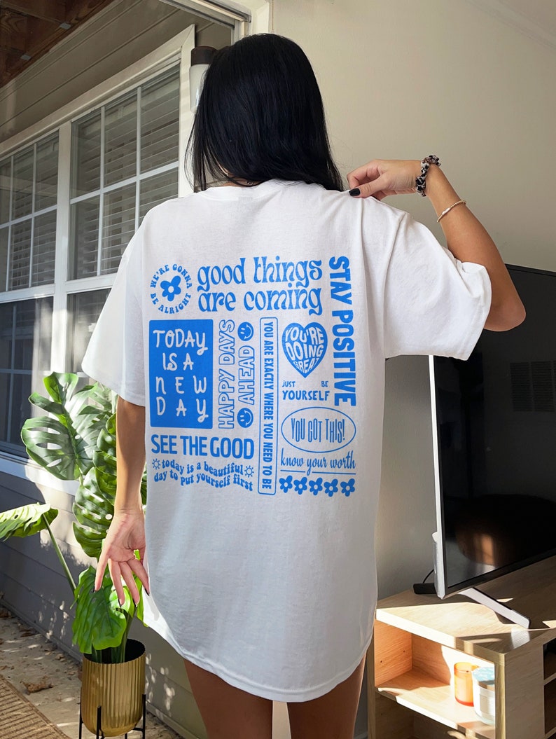 May include: White t-shirt with a blue graphic print of motivational phrases and sayings, including "good things are coming", "stay positive", "happy days ahead", "today is a new day", "see the good", "you got this", and "know your worth".
