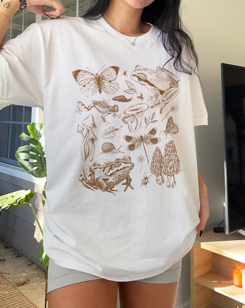 May include: White t-shirt with a brown graphic print of various forest creatures, including butterflies, frogs, mushrooms, and a dragonfly.