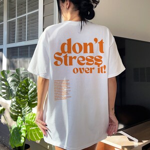 Puede incluir: Camiseta blanca de gran tama&ntilde;o con el texto "don't stress over it!" impreso en naranja en la parte posterior. El texto est&aacute; en una fuente en negrita y retro.