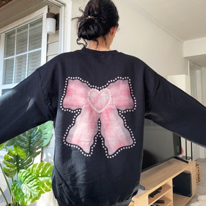 May include: Black sweatshirt with a pink bow and a heart design on the back. The bow is outlined with white dots.