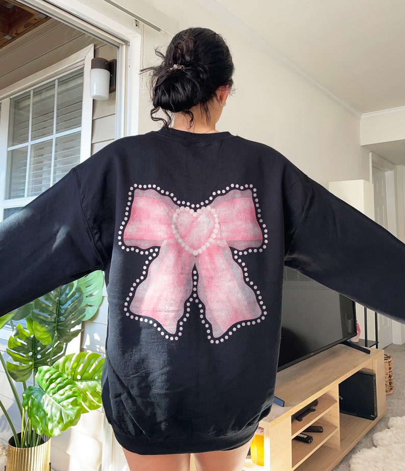 May include: Black sweatshirt with a pink bow and a heart design on the back. The bow is outlined with white dots.