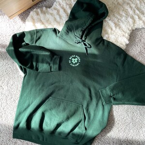 May include: A dark green hoodie with a large pocket on the front. The hoodie has a drawstring hood and a graphic on the front that says "We're gonna be alright" with a flower design.