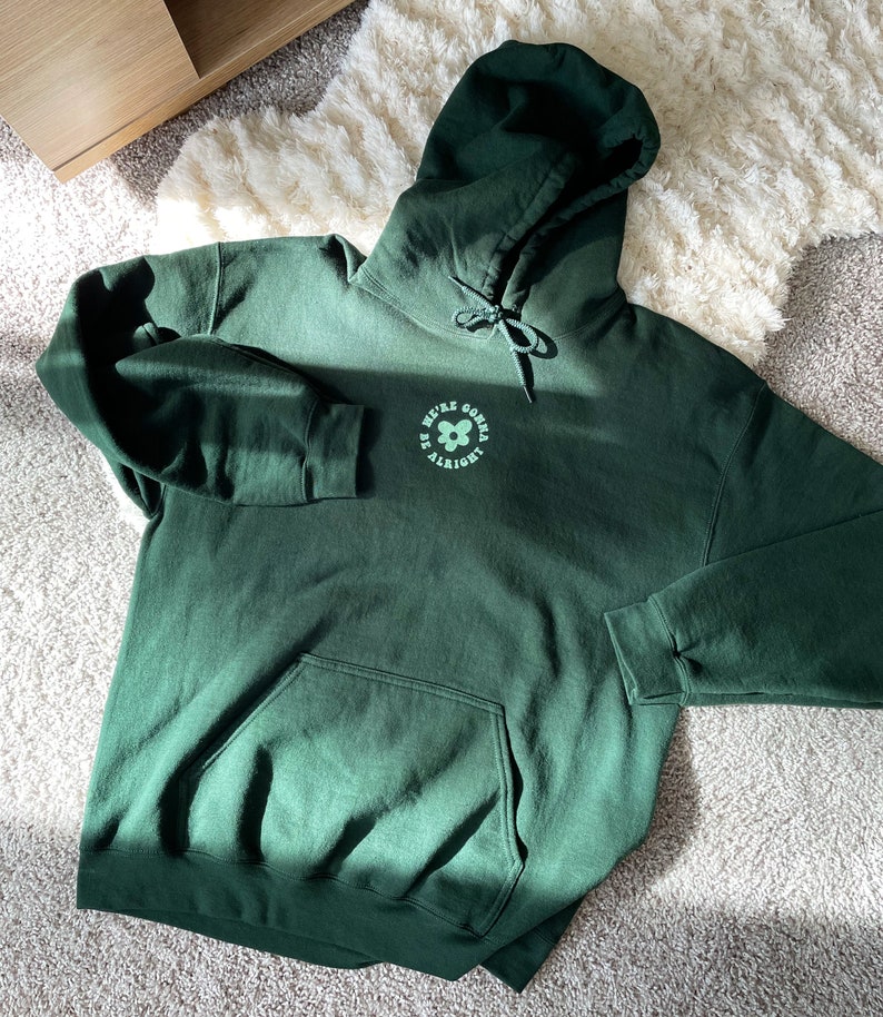 May include: A dark green hoodie with a large pocket on the front. The hoodie has a drawstring hood and a graphic on the front that says "We're gonna be alright" with a flower design.