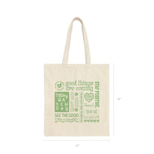 May include: A beige canvas tote bag with green text that reads "good things are coming", "today is a new day", "happy days ahead", "see the good", "you are exactly where you need to be", "you're doing great", "just be yourself", "you got this", "know your worth", and "stay positive". The bag is 15 inches wide and 16 inches tall.