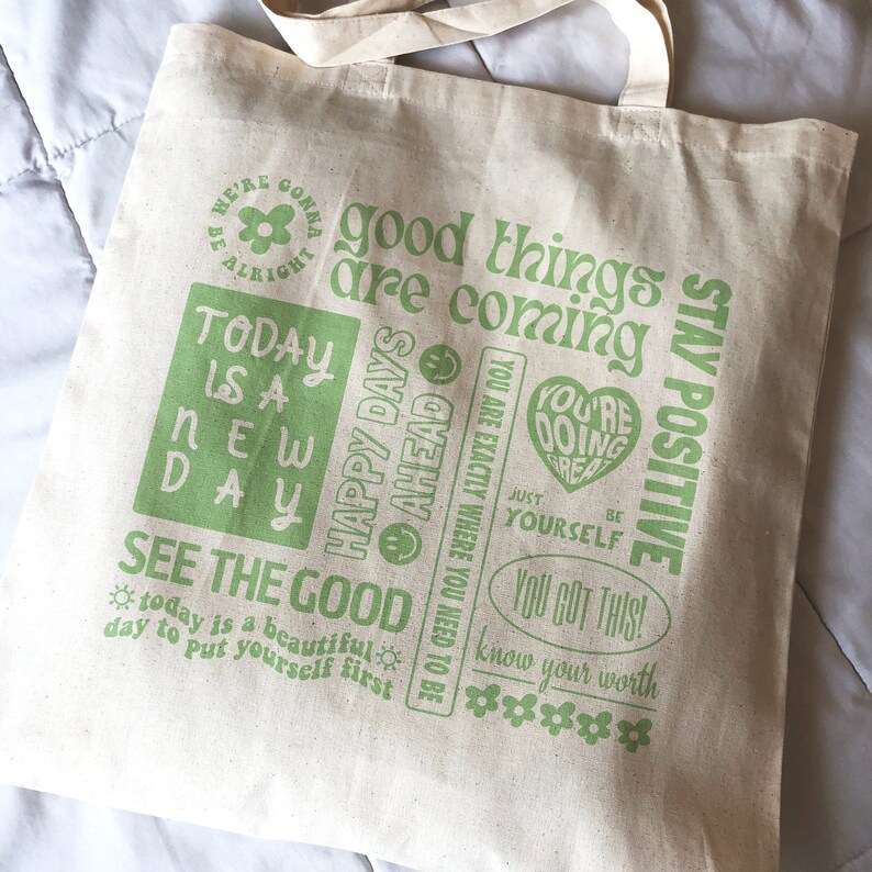 May include: A white canvas tote bag with green text and graphics. The text includes "good things are coming", "stay positive", "today is a new day", "see the good", "you got this", and "know your worth".