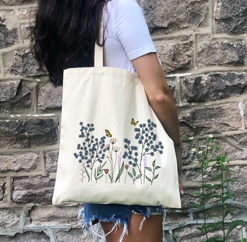 K&ouml;nnte beinhalten: Eine wei&szlig;e Canvas-Tragetasche mit einem Blumendesign mit blauen Blumen, gr&uuml;nen Bl&auml;ttern und orangefarbenen Schmetterlingen. Die Tasche hat einen langen Henkel und ist aus Naturstoff gefertigt.