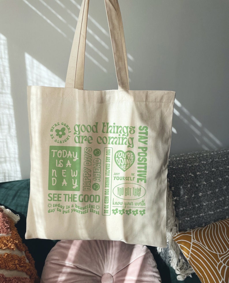 May include: A beige canvas tote bag with green text that reads "good things are coming" and "stay positive". The bag also features other positive affirmations, including "today is a new day", "you're doing great", and "know your worth".