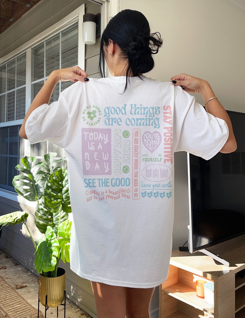 May include: White t-shirt with a colorful graphic print featuring positive affirmations and motivational quotes. The text includes "good things are coming", "today is a new day", "see the good", "stay positive", "happy days", "you are exactly where you need to be", "you got this", "know your worth", and "you're doing just be yourself".
