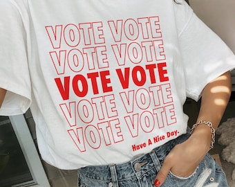 May include: White t-shirt with the phrase "VOTE VOTE" repeated five times in red text. The text is outlined in white. The shirt also has the text "Have A Nice Day." in smaller red text.