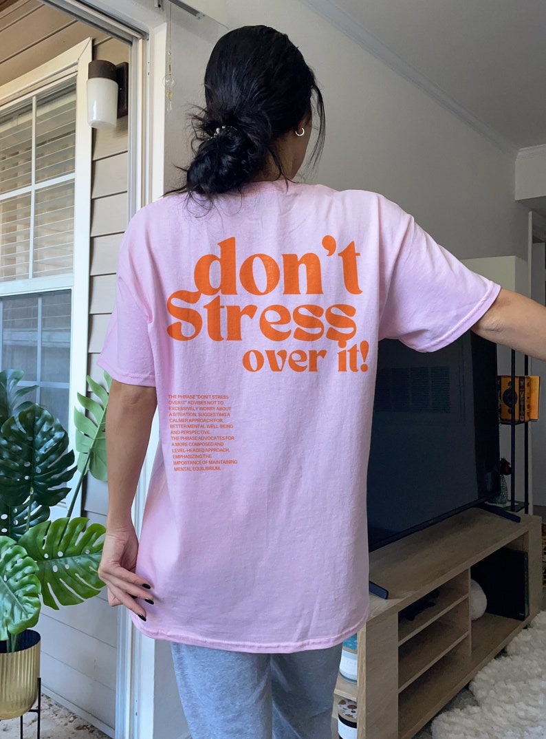 Puede incluir: Camiseta rosa con el texto "don't stress over it!" en naranja. La parte posterior de la camiseta tiene un mensaje sobre la importancia de mantener el equilibrio mental.
