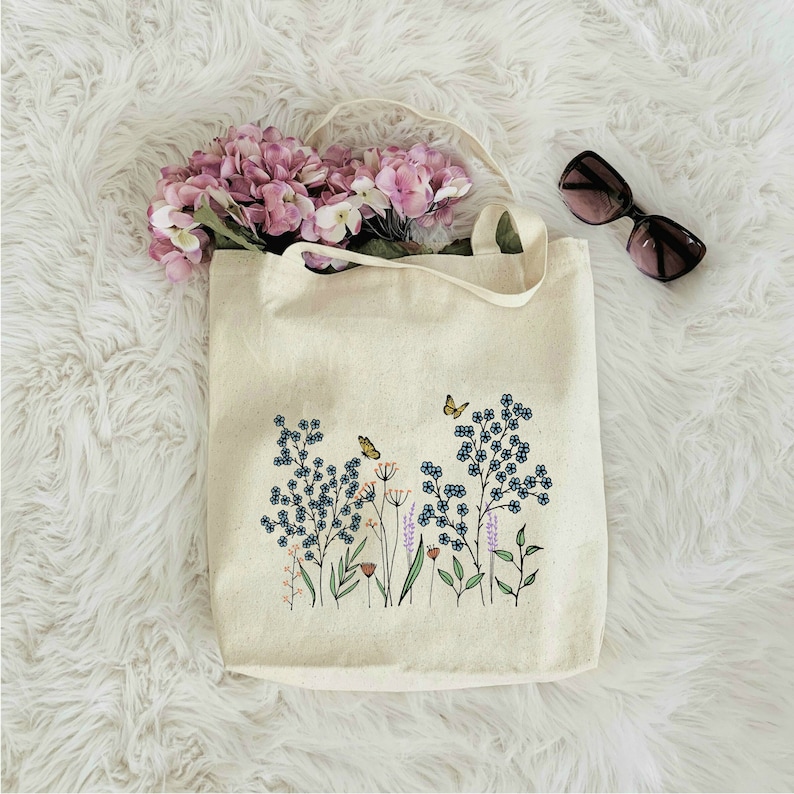 K&ouml;nnte beinhalten: Eine wei&szlig;e Canvas-Tragetasche mit einem Blumendesign mit blauen Blumen, gr&uuml;nen Bl&auml;ttern und orangefarbenen Schmetterlingen. Die Tasche hat eine nat&uuml;rliche Stofftextur und ist perfekt f&uuml;r den Transport von Alltagsgegenst&auml;nden.