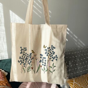 K&ouml;nnte beinhalten: Eine beige Canvas-Tragetasche mit einem Blumendesign mit blauen Blumen, gr&uuml;nen Bl&auml;ttern und einem gelben Schmetterling. Das Design ist mittig auf der Tasche platziert.