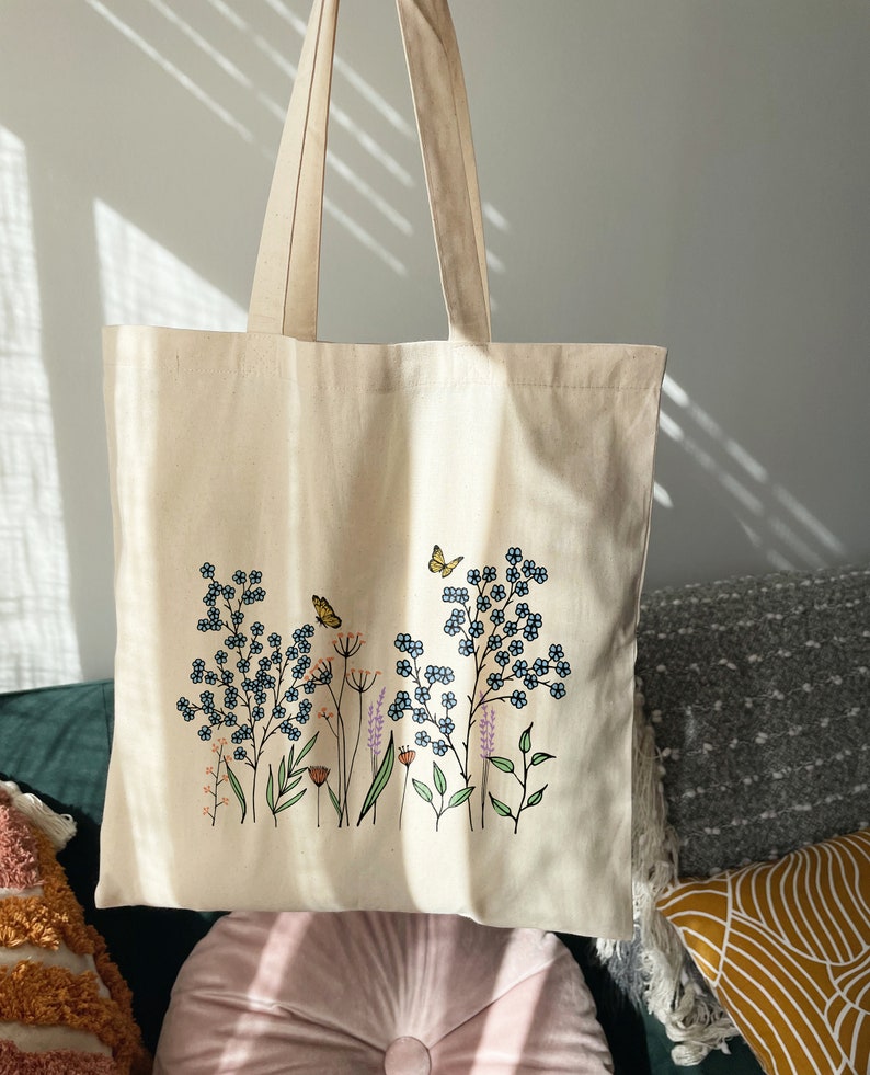 K&ouml;nnte beinhalten: Eine beige Canvas-Tragetasche mit einem Blumendesign mit blauen Blumen, gr&uuml;nen Bl&auml;ttern und einem gelben Schmetterling. Das Design ist mittig auf der Tasche platziert.