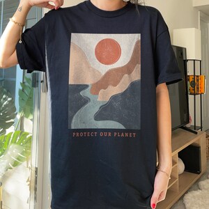 May include: Black t-shirt with a graphic print of a stylized landscape with a river, mountains, and a red sun. The text "PROTECT OUR PLANET" is printed below the graphic.