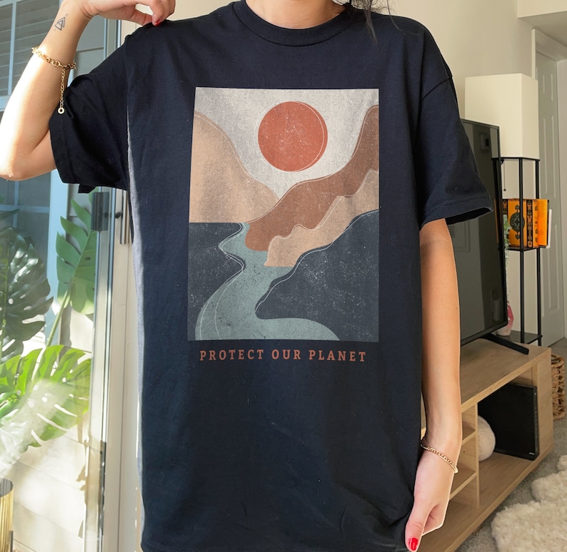 May include: Black t-shirt with a graphic print of a stylized landscape with a river, mountains, and a red sun. The text "PROTECT OUR PLANET" is printed below the graphic.