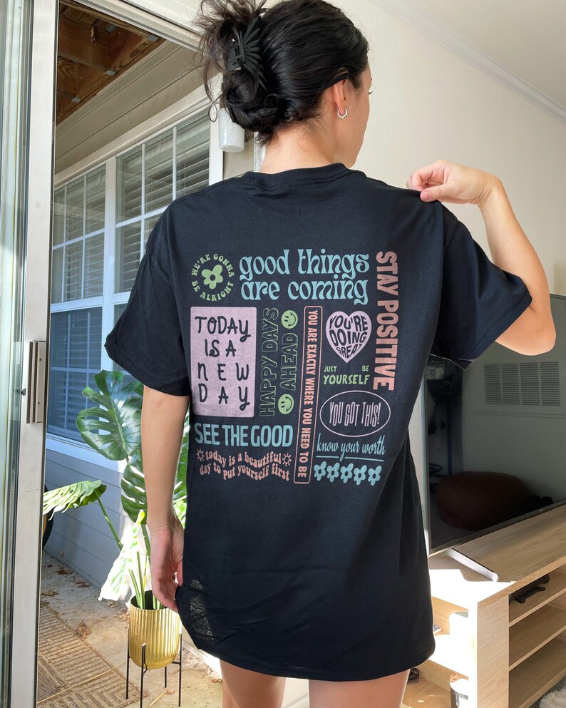 May include: Black t-shirt with a colorful graphic print featuring positive affirmations and motivational quotes. The text includes "good things are coming", "stay positive", "happy days ahead", "see the good", "today is a new day", "you're doing great", "you got this", and "know your worth".