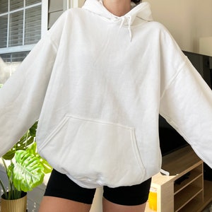 May include: A white hooded sweatshirt with a kangaroo pocket. The sweatshirt is made of a soft material and has a drawstring at the hood. The sleeves are long and the fit is oversized. The person is wearing black shorts.