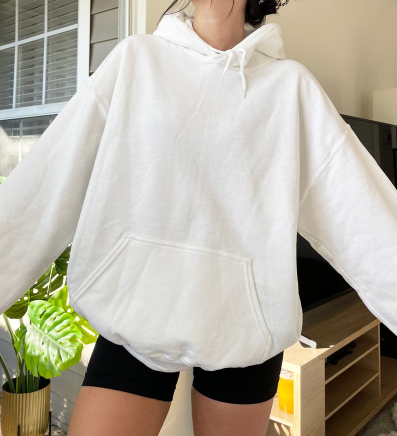 May include: A white hooded sweatshirt with a kangaroo pocket. The sweatshirt is made of a soft material and has a drawstring at the hood. The sleeves are long and the fit is oversized. The person is wearing black shorts.