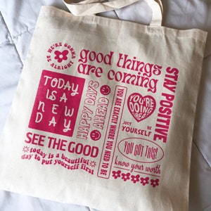 May include: A white canvas tote bag with pink text that reads "We're gonna be alright", "good things are coming", "Today is a new day", "Happy days ahead", "You are exactly where you need to be", "You're doing great", "Just be yourself", "You got this!", "Know your worth", and "Stay positive".