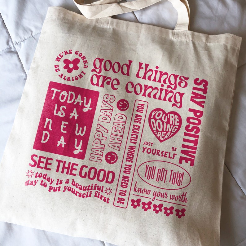 May include: A white canvas tote bag with pink text that reads "We're gonna be alright", "good things are coming", "Today is a new day", "Happy days ahead", "You are exactly where you need to be", "You're doing great", "Just be yourself", "You got this!", "Know your worth", and "Stay positive".