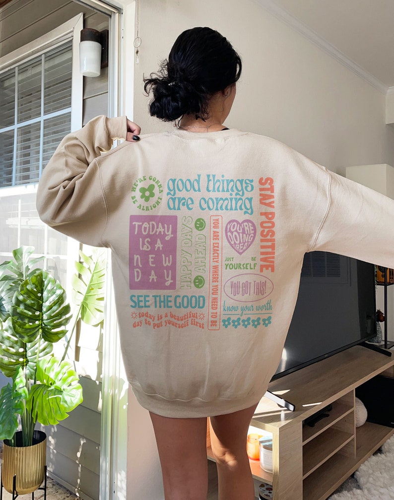 May include: A beige sweatshirt with a colorful graphic print featuring positive affirmations and motivational quotes. The text includes "good things are coming", "stay positive", "today is a new day", "see the good", and "you got this!"