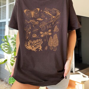 May include: A brown t-shirt with a graphic print of various forest creatures, including frogs, butterflies, dragonflies, mushrooms, and snails. The print is in a light brown color.