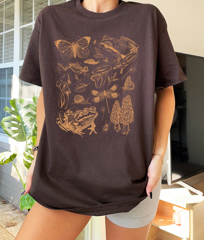 May include: A brown t-shirt with a graphic print of various forest creatures, including frogs, butterflies, dragonflies, mushrooms, and snails. The print is in a light brown color.