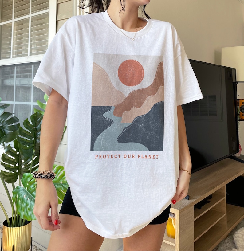 May include: White oversized t-shirt with a graphic print of a sunset over mountains and a river. The text "PROTECT OUR PLANET" is printed below the graphic.