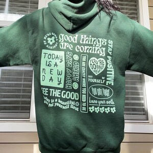 May include: Green hooded sweatshirt with a variety of positive messages printed on the back in a light green color. The text includes "good things are coming", "today is a new day", "happy days ahead", "you're doing great", "see the good", "you got this", and "know your worth".
