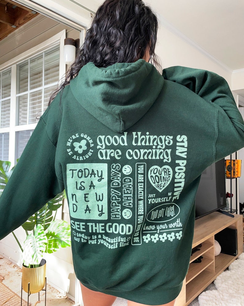 May include: Green hooded sweatshirt with a variety of positive messages printed on the back in a light green color. The text includes "good things are coming", "today is a new day", "happy days ahead", "stay positive", "see the good", "you're doing great", "just be yourself", "you got this", and "know your worth".