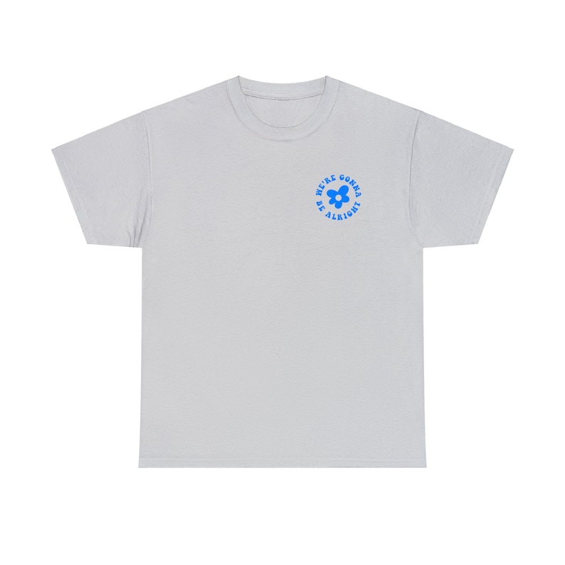 May include: A light gray t-shirt with a blue flower graphic and the text "We're gonna be alright" in a circle around the flower.