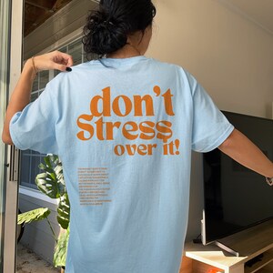 Puede incluir: Camiseta azul claro con el texto "don't stress over it!" impreso en naranja en la parte posterior. El texto est&aacute; en una fuente retro en negrita. La frase "don't stress over it" es un recordatorio para mantener la calma y la relajaci&oacute;n.