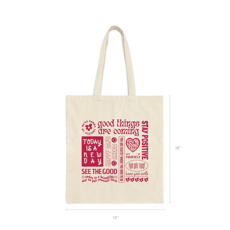 May include: A beige canvas tote bag with red text that reads "good things are coming", "today is a new day", "happy days ahead", "see the good", "you are exactly where you need to be", "you're doing great", "just be yourself", "you got this", "know your worth", and "stay positive". The bag measures 15 inches wide by 16 inches tall.
