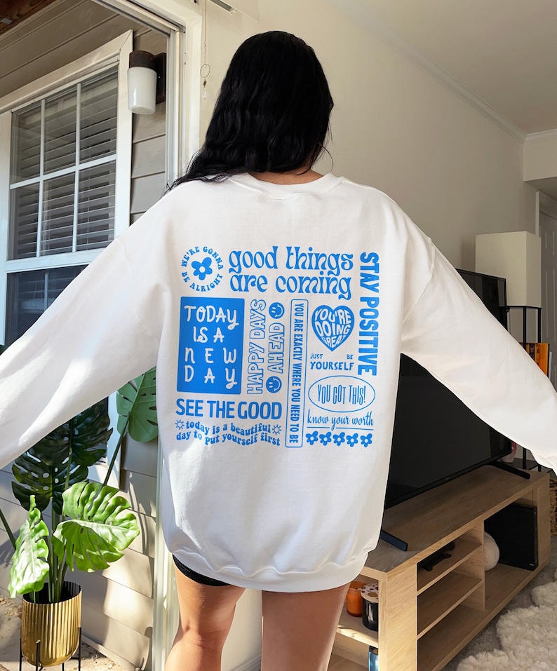 May include: White sweatshirt with blue text that says "good things are coming", "stay positive", "today is a new day", "happy days ahead", "you are exactly where you need to be", "you're doing great", "just be yourself", "you got this", "know your worth", and "see the good".