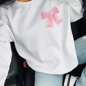 May include: White sweatshirt with a pink watercolor bow design. The bow has a pearl-like outline.