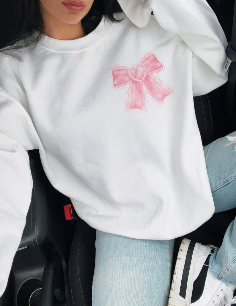May include: White sweatshirt with a pink watercolor bow design. The bow has a pearl-like outline.