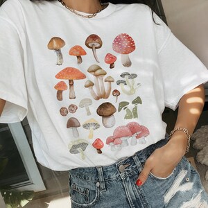 May include: White t-shirt with a colorful mushroom print. The mushrooms are various shapes and sizes and have red, brown, white, and green accents.