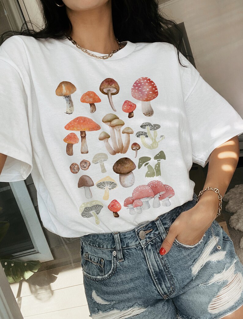 May include: White t-shirt with a colorful mushroom print. The mushrooms are various shapes and sizes and have red, brown, white, and green accents.