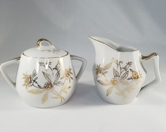 May include: A white porcelain sugar bowl and creamer set with gold floral designs. The set has a lid and two handles on the sugar bowl.