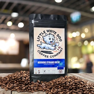 May include: A black coffee bag with the text "Little White Dog Coffee Company" and a cartoon dog logo. The bag is filled with coffee beans and the text "Morning Dynamo Brew" is visible. The bag is sitting on a wooden surface.
