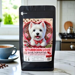 May include: Black coffee bag with a heart-shaped design featuring a white dog wearing a red bow tie. The bag reads "STUBBORN LOVE" and "Specialty Small Batch Coffee". A blue coffee cup and saucer are in the foreground.