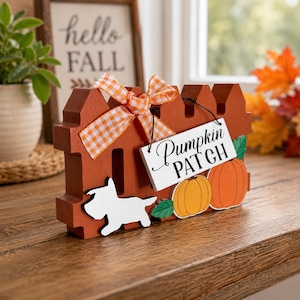 May include: A burnt orange wooden picket fence decoration featuring a white silhouette of a dog, two orange pumpkins with green leaves, and a white rectangular sign with the black text Pumpkin PATCH, accented by an orange and white gingham bow.