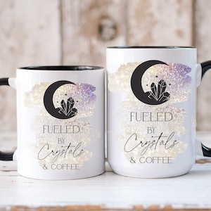 K&ouml;nnte beinhalten: Zwei wei&szlig;e Keramikbecher mit schwarzen R&auml;ndern und Griffen. Die Becher haben ein Aquarell-Design mit einer Mondsichel und Kristallen. Der Text auf den Bechern lautet "Fueled by Crystals & Coffee".