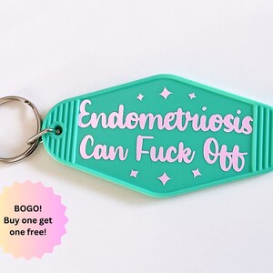 May include: A teal motel-style keychain with a silver metal ring. The keychain features the text "Endometriosis Can Fuck Off" in light pink script lettering, accented by four small pink star shapes above and below the text.