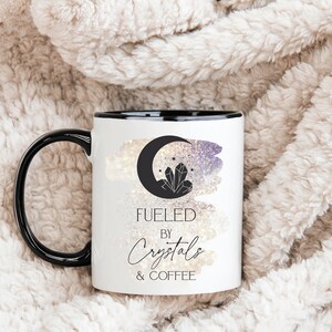K&ouml;nnte beinhalten: Eine wei&szlig;e Keramik-Kaffeetasse mit schwarzem Rand und Henkel. Die Tasse zeigt eine Schwarzwei&szlig;illustration eines zunehmenden Mondes mit Kristallen und Sternen. Der Text "Fueled by Crystals & Coffee" ist in Schwarz unter der Illustration aufgedruckt.