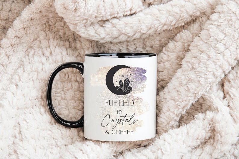 K&ouml;nnte beinhalten: Eine wei&szlig;e Keramik-Kaffeetasse mit schwarzem Rand und Henkel. Die Tasse zeigt eine Schwarzwei&szlig;illustration eines zunehmenden Mondes mit Kristallen und Sternen. Der Text "Fueled by Crystals & Coffee" ist in Schwarz unter der Illustration aufgedruckt.